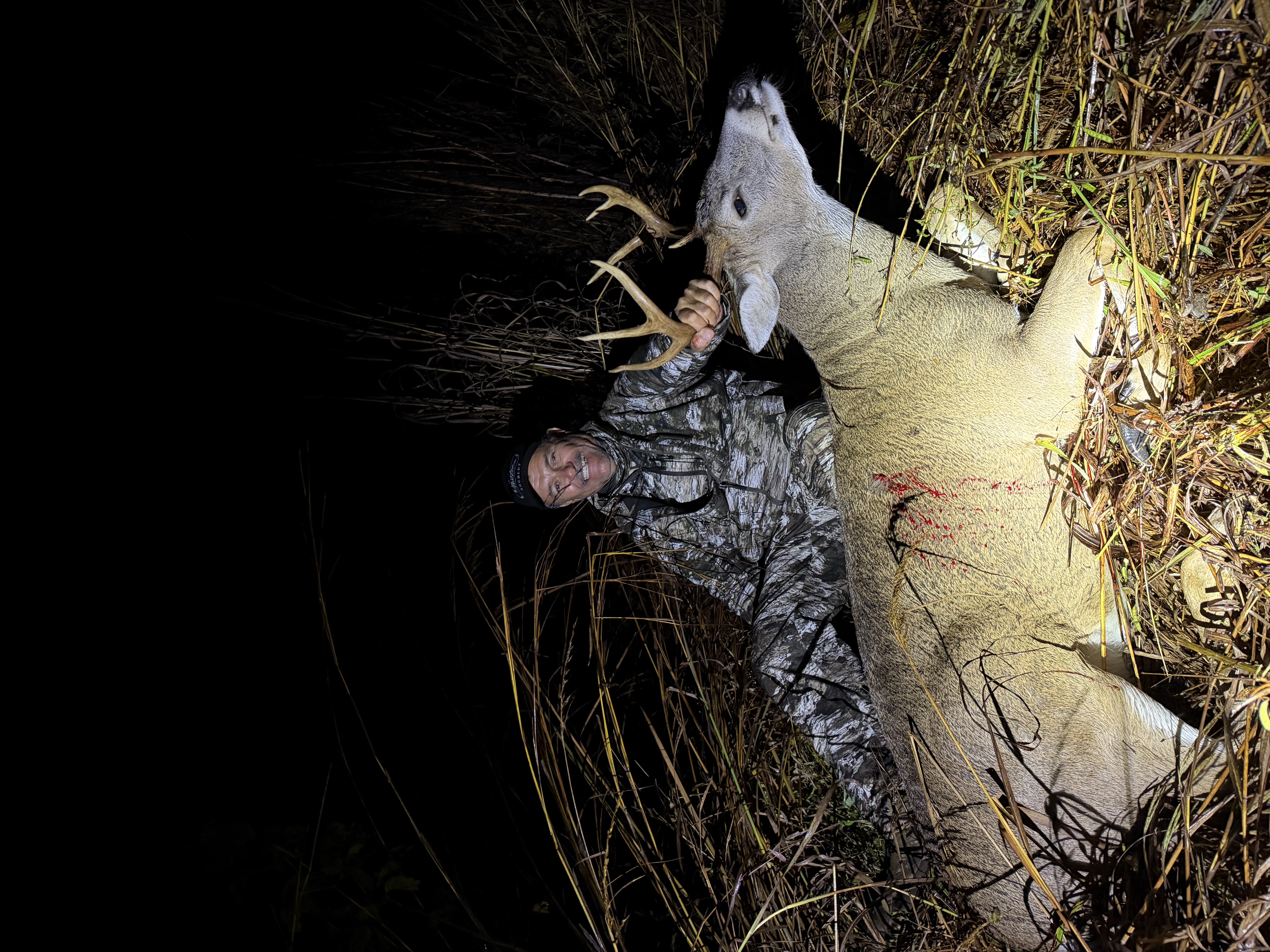 Hunter in camouflage with harvested whitetail buck in tall grass at night