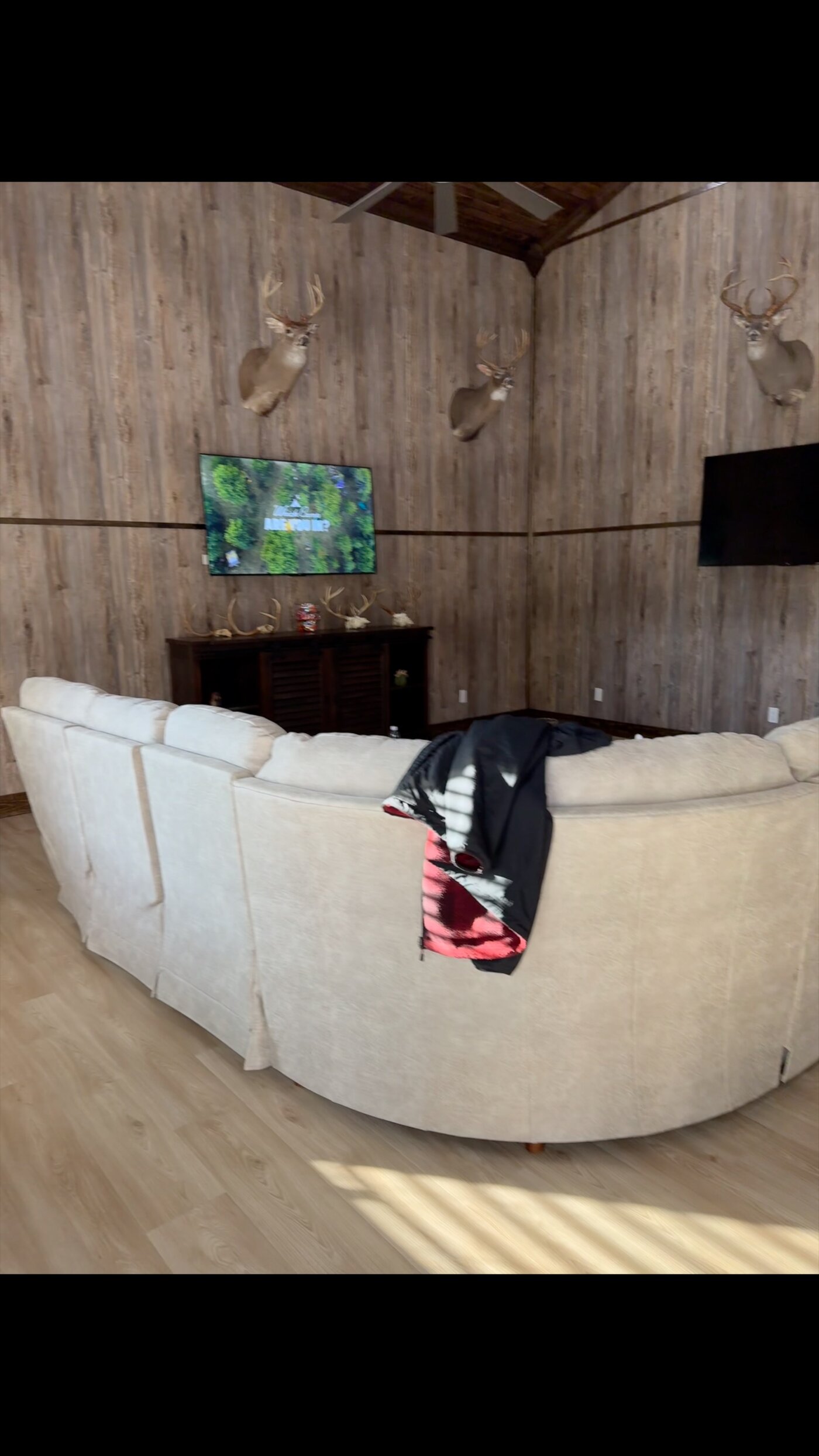 Spacious lodge lounge with a curved sectional sofa, trophy whitetail mounts, and dual flat-screen televisions